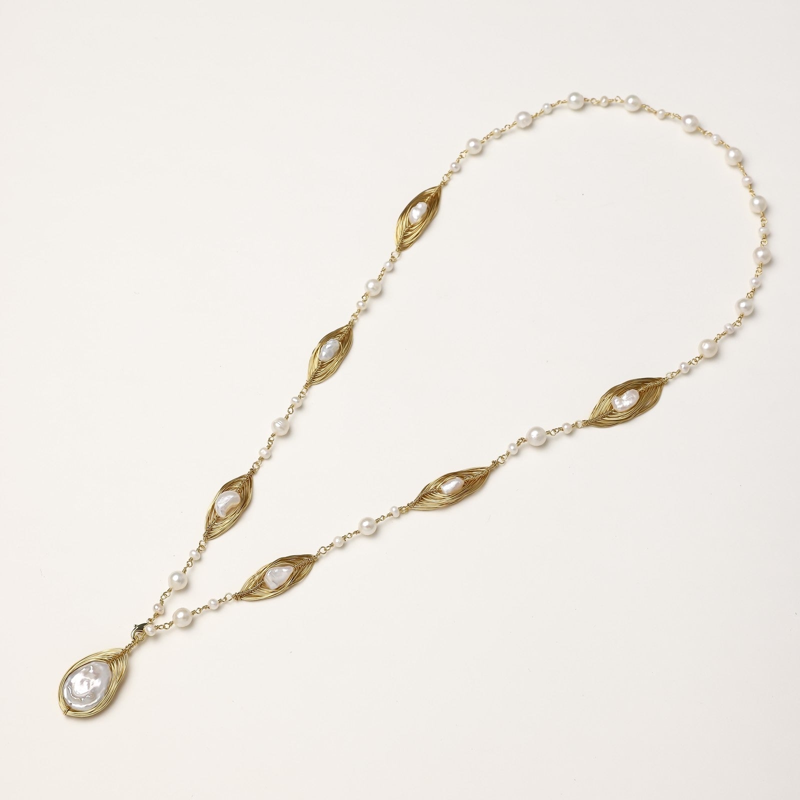 Long gold-plated necklace with eye-shaped elements, freshwater pearls, and baroque pearl pendant on neutral background.