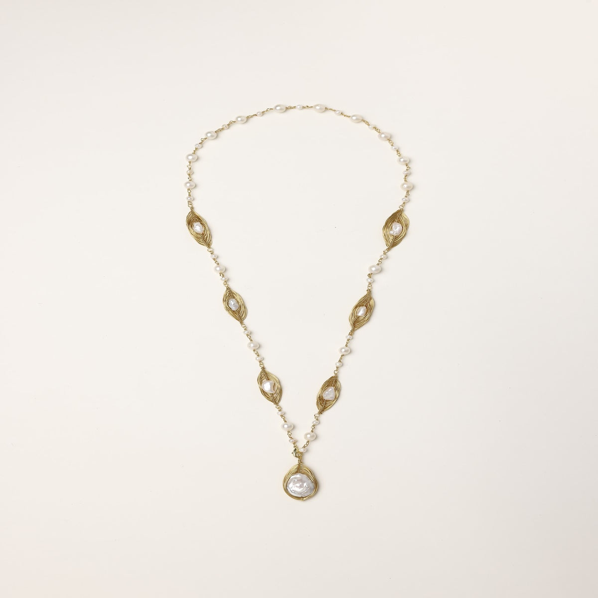 Long gold-plated necklace with eye-shaped elements, freshwater pearls, and baroque pearl pendant on neutral background.