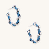 Twisted hoop earrings featuring blue denim fabric braided with gold-plated metal in a spiral design on white background.