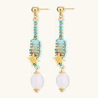 Gold-plated dangle earrings with turquoise threading, star charms, and white pearl beads against cream background.