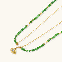Layered necklace with green stone beads, gold spacers, white accents, and protective eye pendant on cream background.