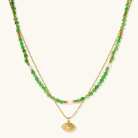 Layered necklace with green stone beads, gold spacers, white accents, and protective eye pendant on cream background.