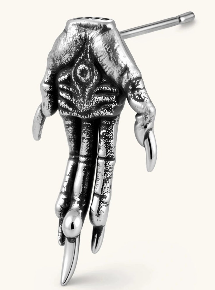 Silver stainless steel stud earring shaped like a skeletal demon claw hand with an eye motif on the palm and sharp talon fingers.